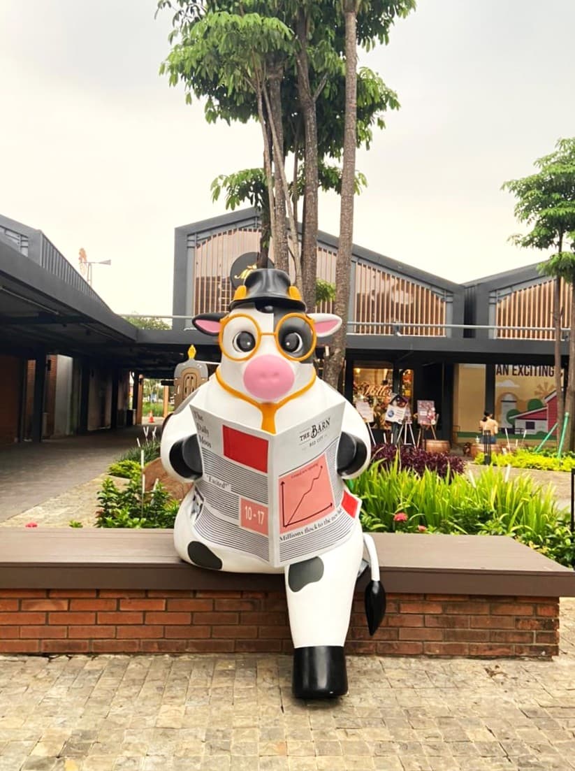 Model lembu gergasi bercermin mata dan bertopi sedang membaca surat khabar di atas bangku di The Barn, BSD City, Indonesia